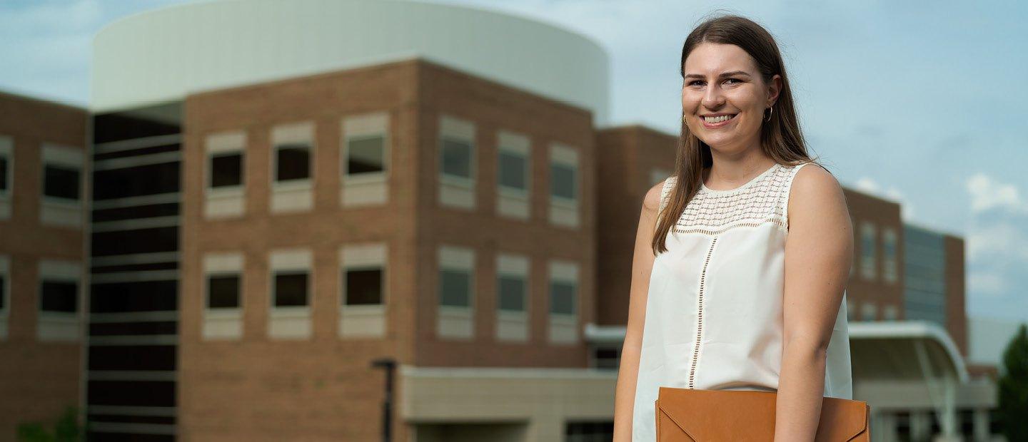 A woman holding a brown folder standing in front of a brown building at Oakland University.