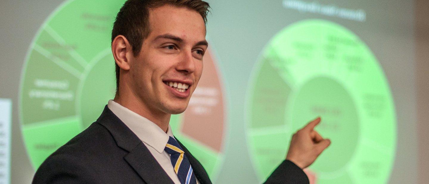 A young man in a suit, pointing to a neon green chart on a screen behind him.